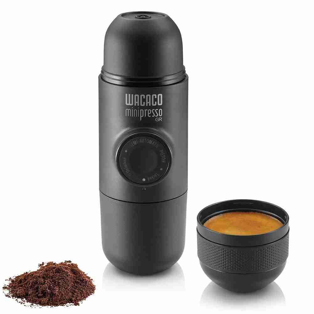 The Wacaco Minipresso portable espresso maker next to a freshly brewed cup of coffee and a freshly ground pile of coffee grounds. The espresso maker for best coffee maker for camping. 