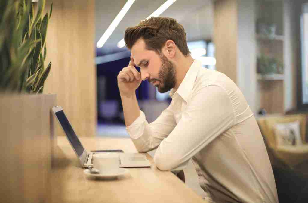 Man in white collared shirt working on laptop holding his head